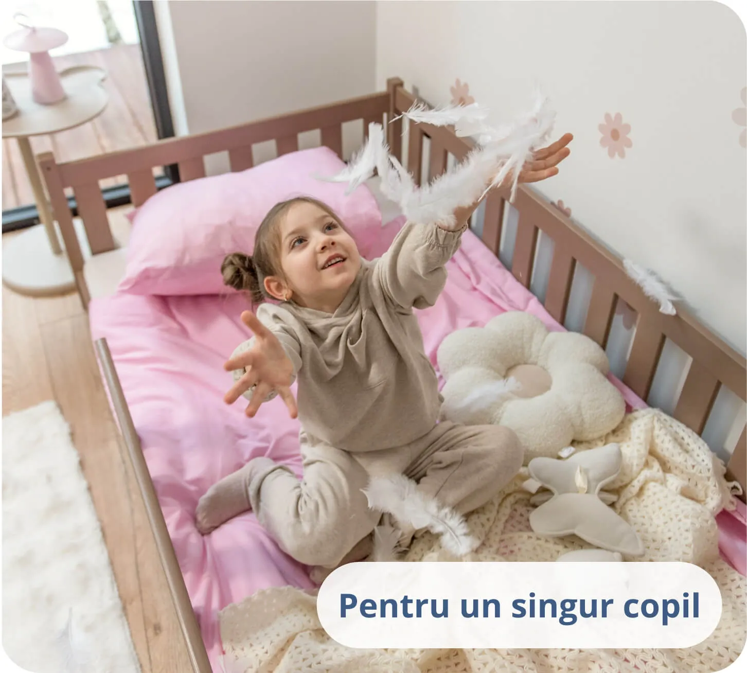 Fetiță jucându-se cu pene albe pe lenjerie roz într-un pat de culoarea ciocolatei.